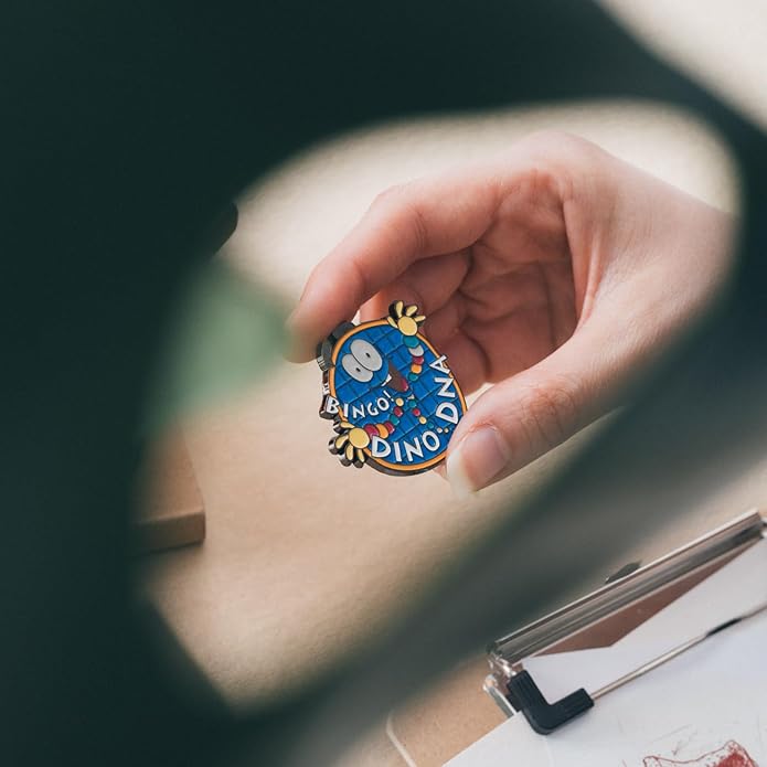 Hand holding a colorful enamel pin with 'Bingo Dino DNA' branding through a magnifying glass.