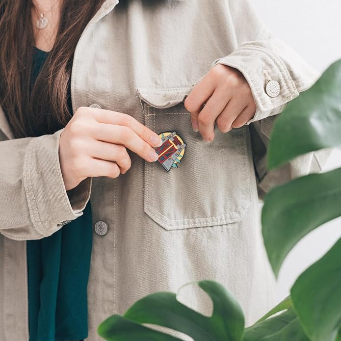 Person wearing a beige jacket with a colorful brooch, surrounded by green leaves.
